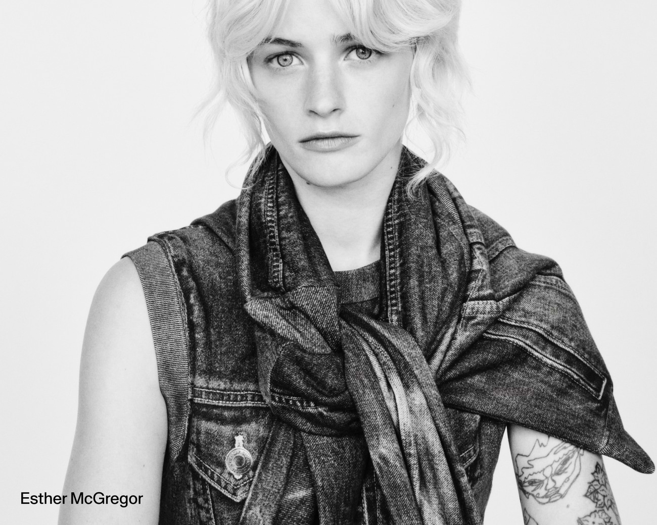 Black-and-white portrait of a person with short light hair, wearing a sleeveless denim vest with a denim jacket tied at the shoulders and a tattooed forearm.