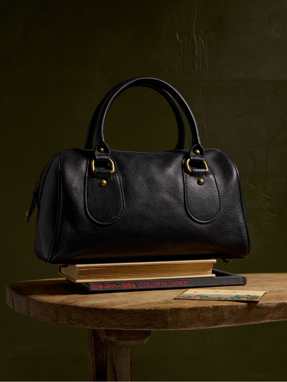 Black pebbled leather handbag with brass hardware and rounded handles, resting on a stack of books on a wooden table.