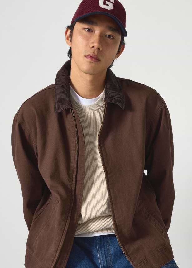 Person in a maroon baseball cap with a “G,” brown jacket over a cream sweater and white tee, and blue jeans, against a plain background.