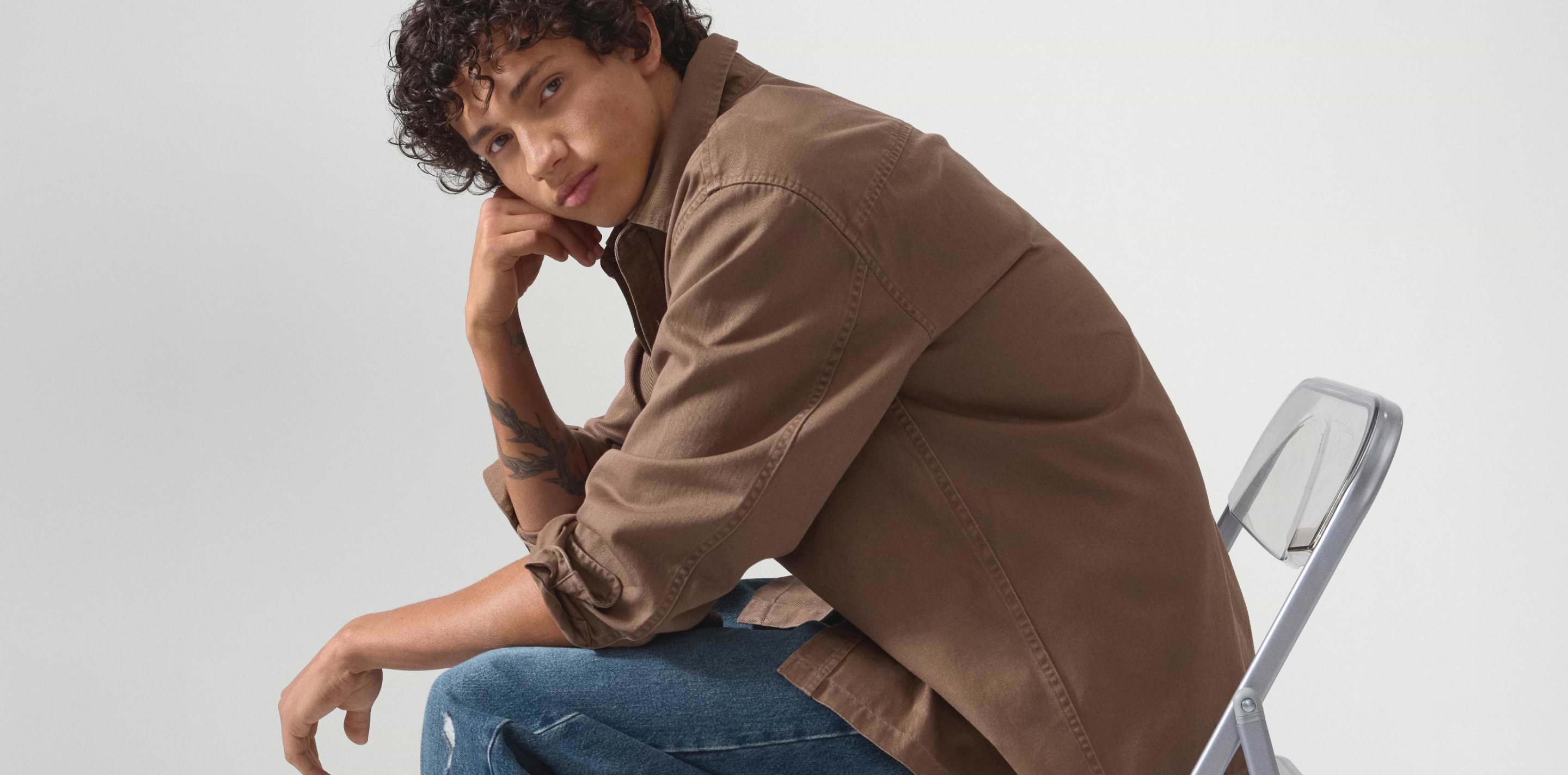Person with curly hair sits sideways on a metal chair, wearing a brown shirt and blue jeans, resting their chin on their hand.