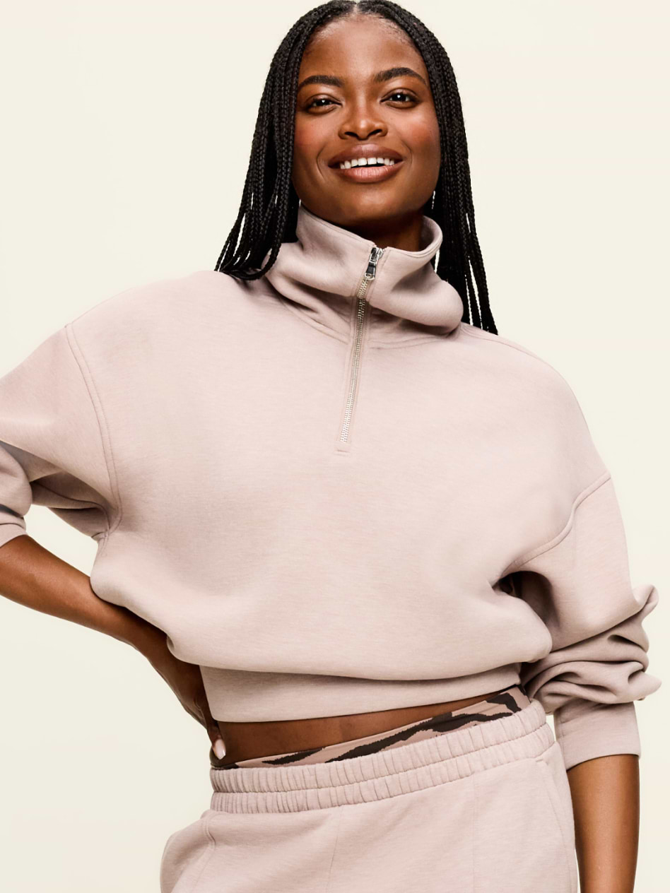 Smiling person with long braids wearing a light beige half‑zip mock‑neck cropped sweatshirt and matching joggers.