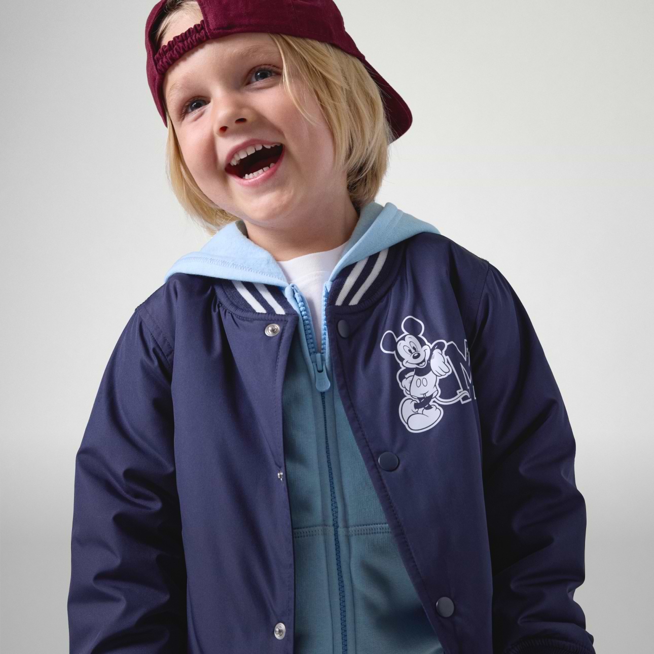 Smiling child wearing a backwards burgundy cap and a navy jacket with a Mickey Mouse graphic over a light blue hoodie.