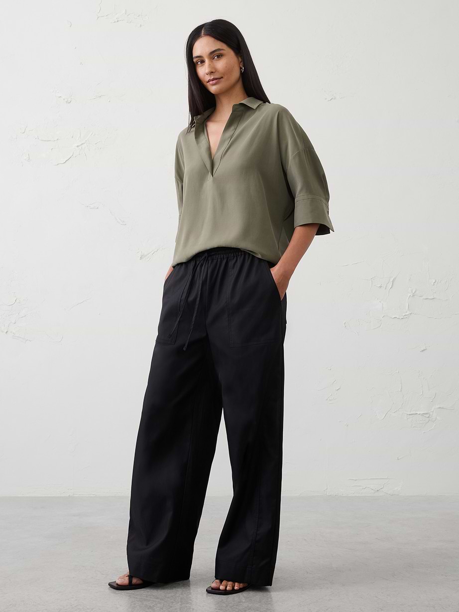 Person standing against a plain wall, wearing an olive green V-neck collared blouse, black wide-leg drawstring pants, and black sandals.
