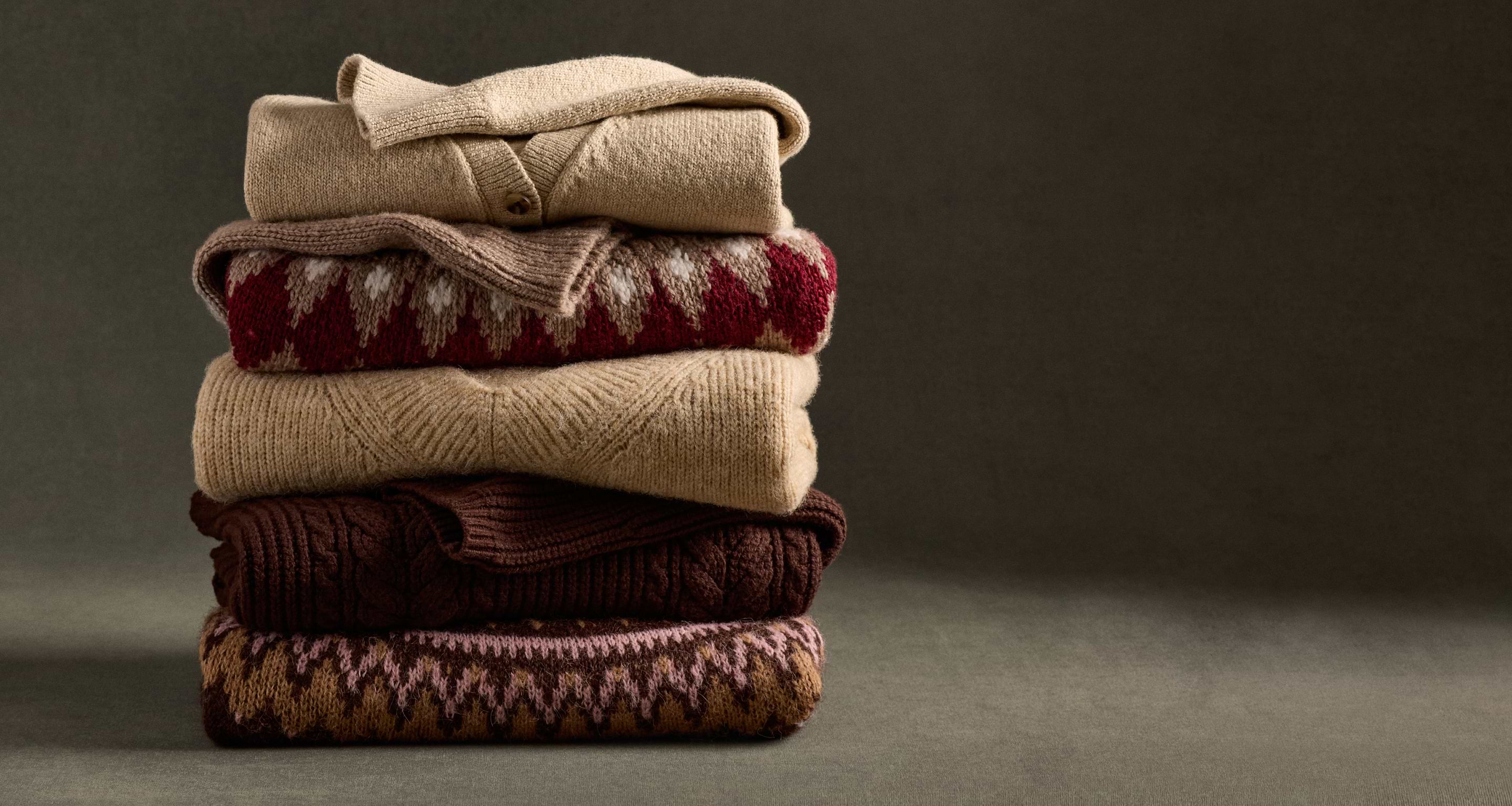 Stack of five folded knit sweaters in beige, brown, and red tones, some with patterns, against a dark background.
