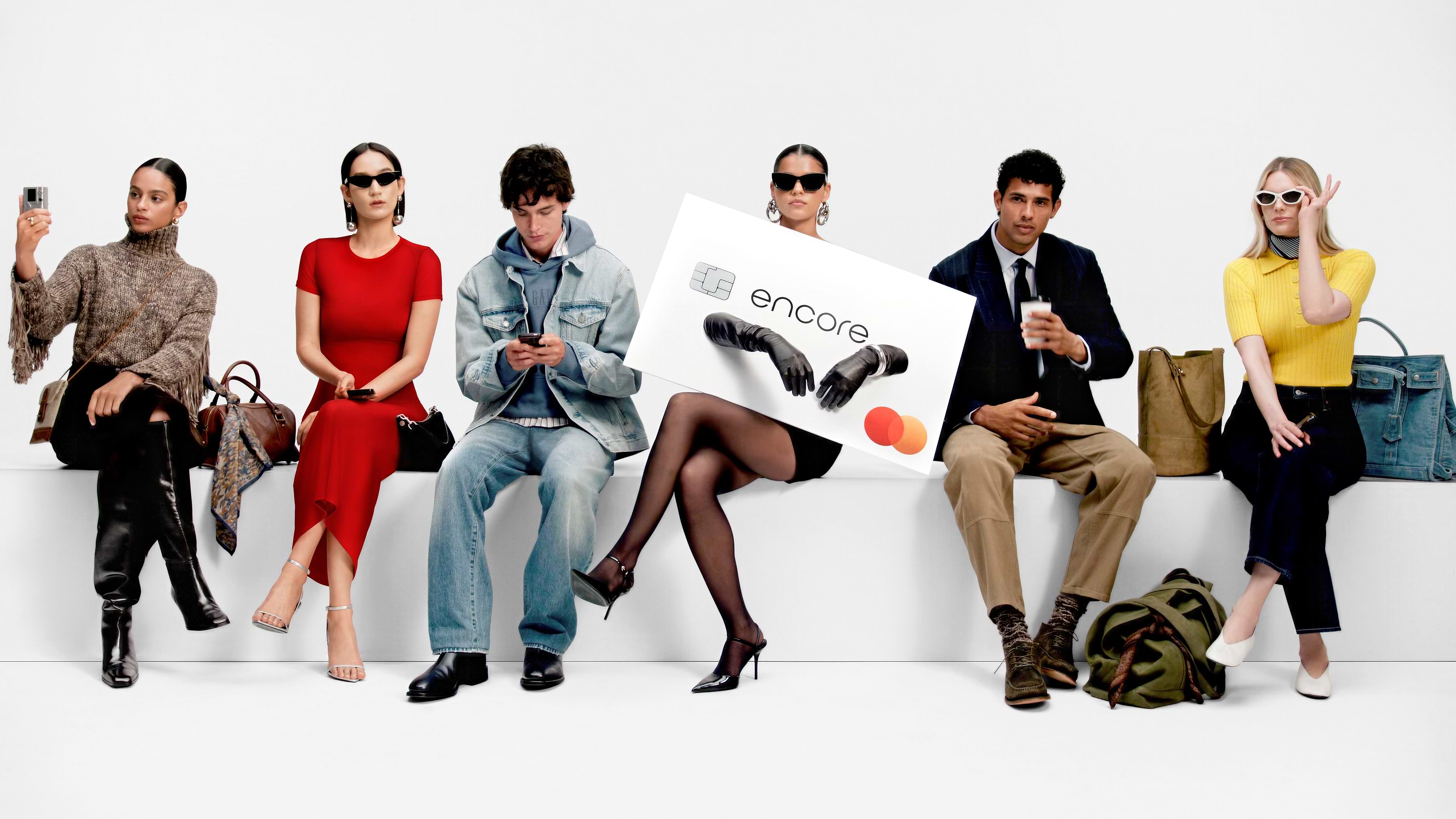 Six stylish people sit on a white bench; the center person in sunglasses holds an oversized “encore” credit card with a Mastercard logo, with bags by their feet.