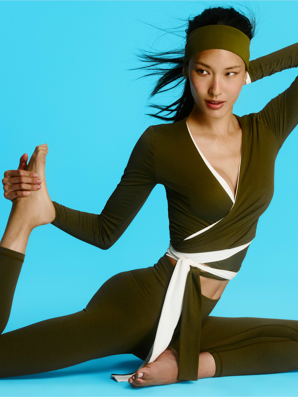 Person in an olive-green wrap top and leggings with a white sash and headband sits in a yoga pose, holding one foot behind them against a bright blue background.