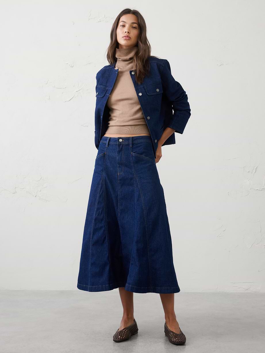 Person wearing a navy denim jacket and matching A-line midi skirt with a tan turtleneck and brown flats, standing indoors.
