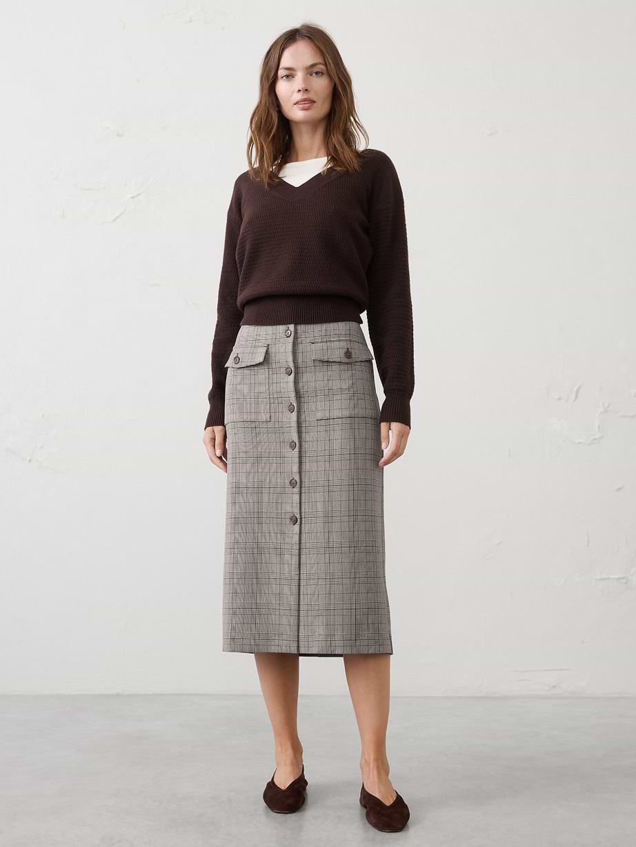 Person standing against a plain wall wearing a dark brown V-neck sweater over a white top, a gray plaid button-front midi skirt with flap pockets, and brown flats.