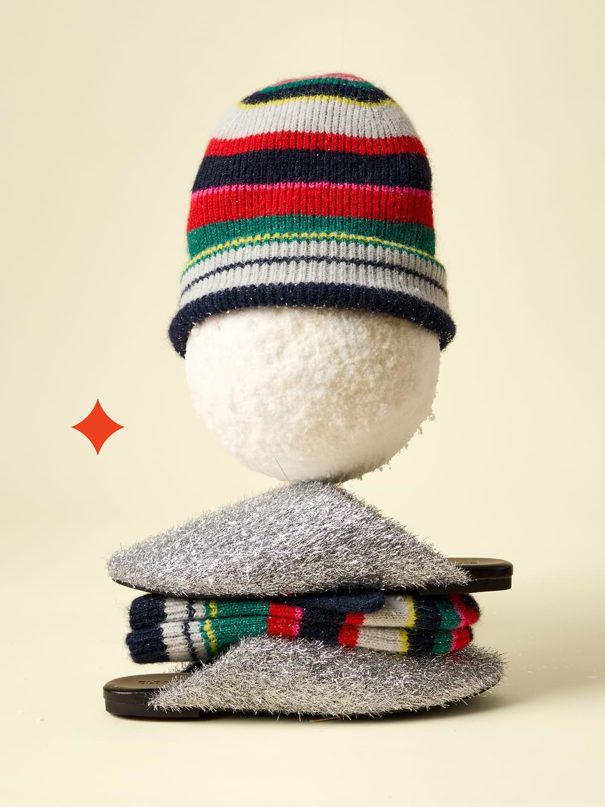 Snowball “head” in a colorful striped beanie balanced on silver fuzzy slippers with folded striped socks, cream background.