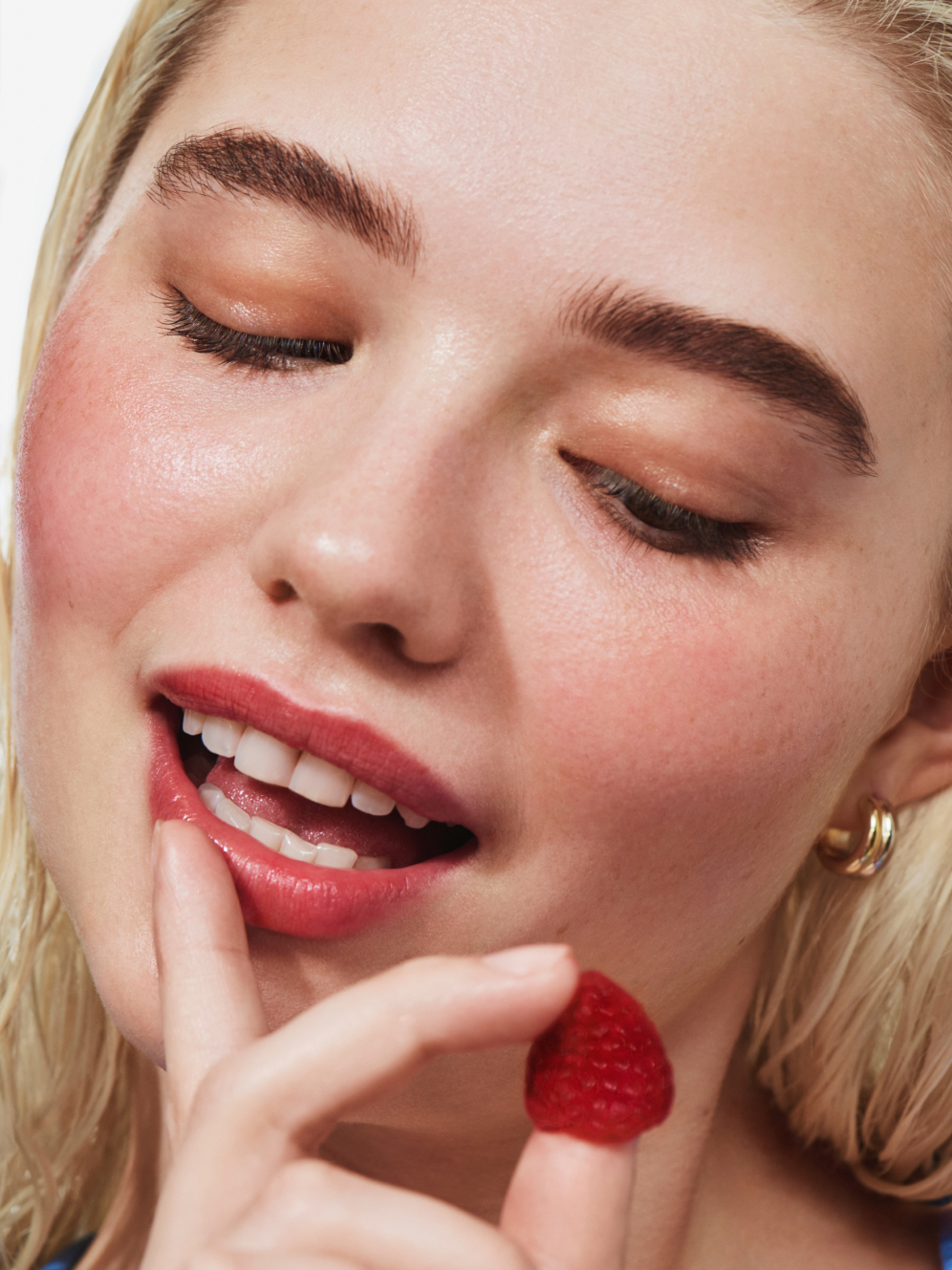 Close-up of a person with closed eyes and glossy red lips, holding a raspberry near their mouth and touching their lip.
