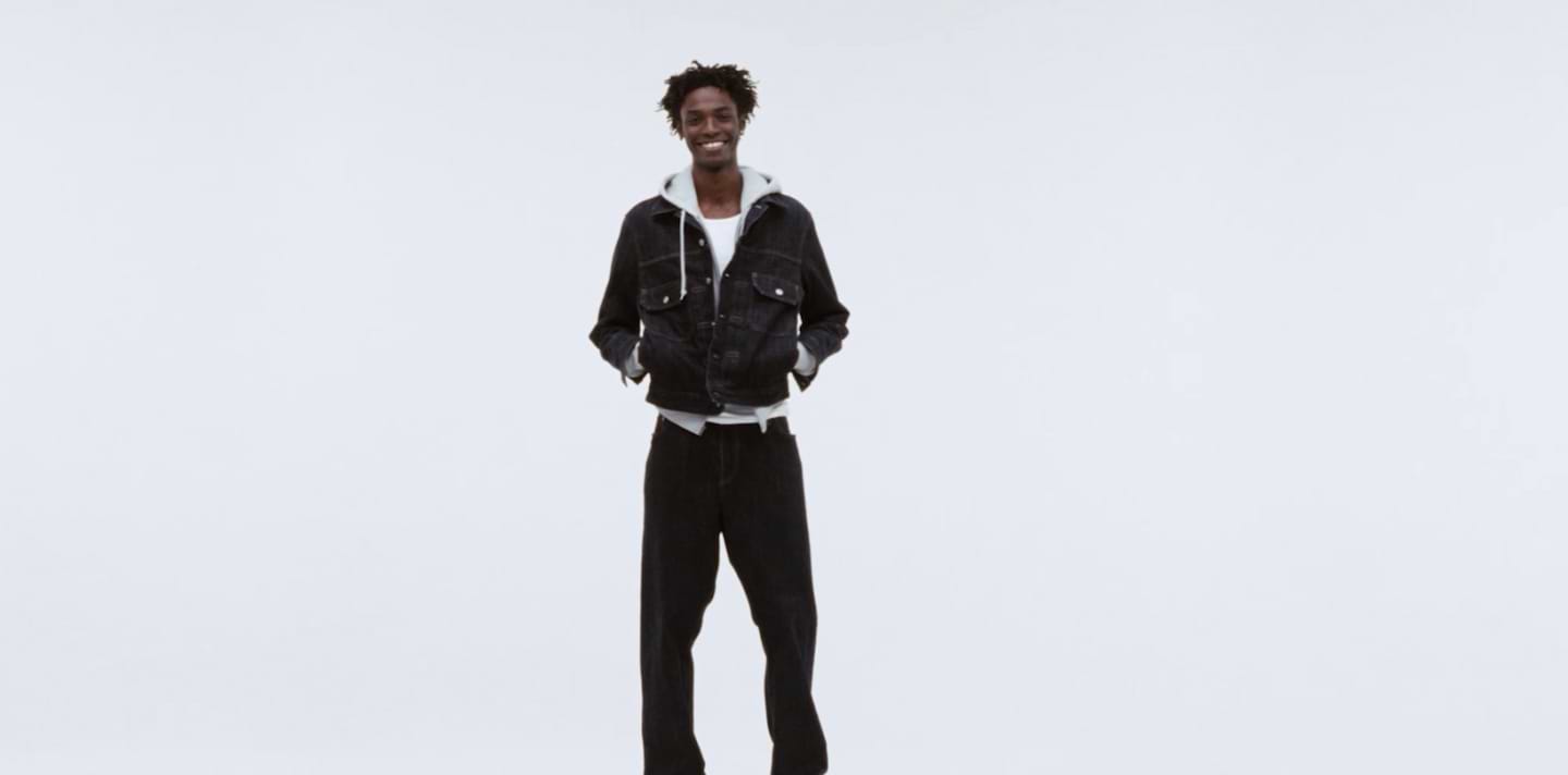 Smiling person in a dark denim jacket over a gray hoodie, hands in pockets, on a plain white backdrop.