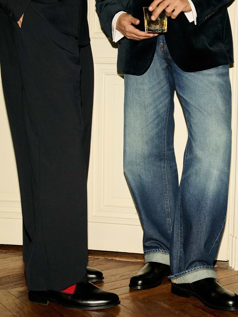 Two people standing indoors from waist down: one in black suit trousers with red socks and black shoes, the other in faded jeans and a dark blazer holding a glass of amber drink; both on a wooden floor.