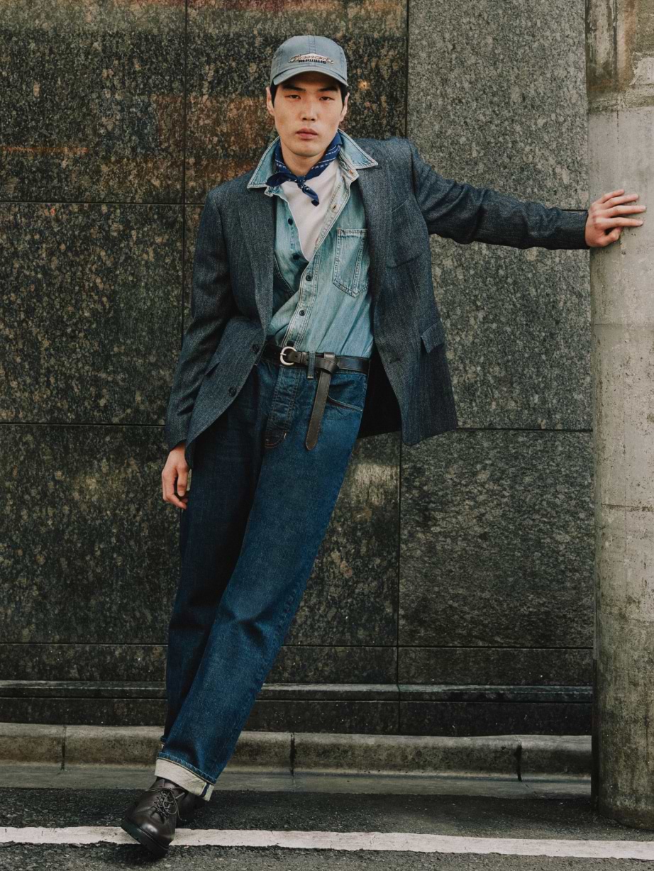Person in jeans, denim shirt, and dark blazer, wearing a cap and neck scarf, leans against a stone wall.