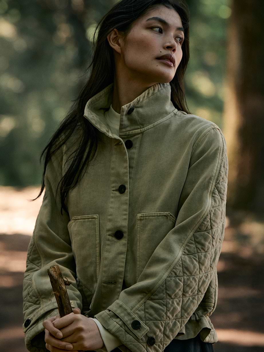 Person in a forest wearing an olive quilted jacket, holding a stick and looking off to the side.