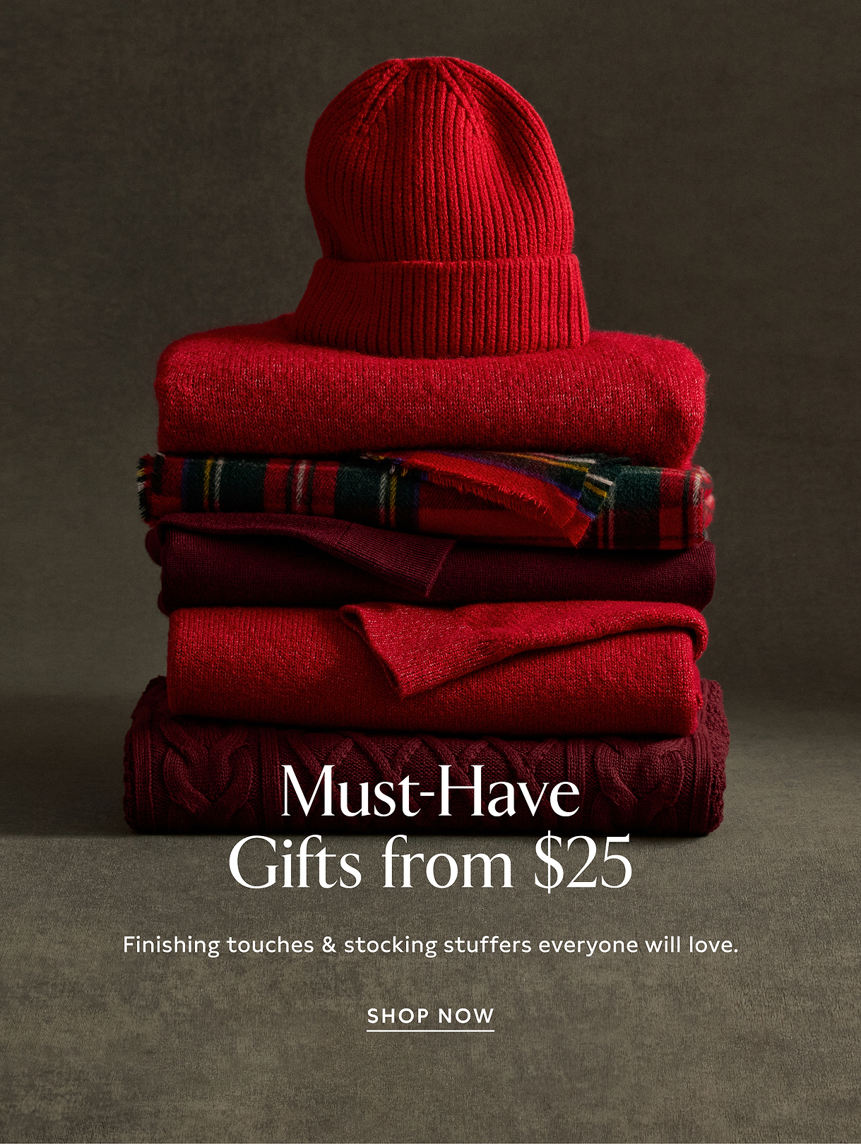 Stack of red winter knitwear—beanie, sweaters, and a plaid scarf—on a dark background with overlaid text: “Must‑Have Gifts from $25” and “Shop Now.”