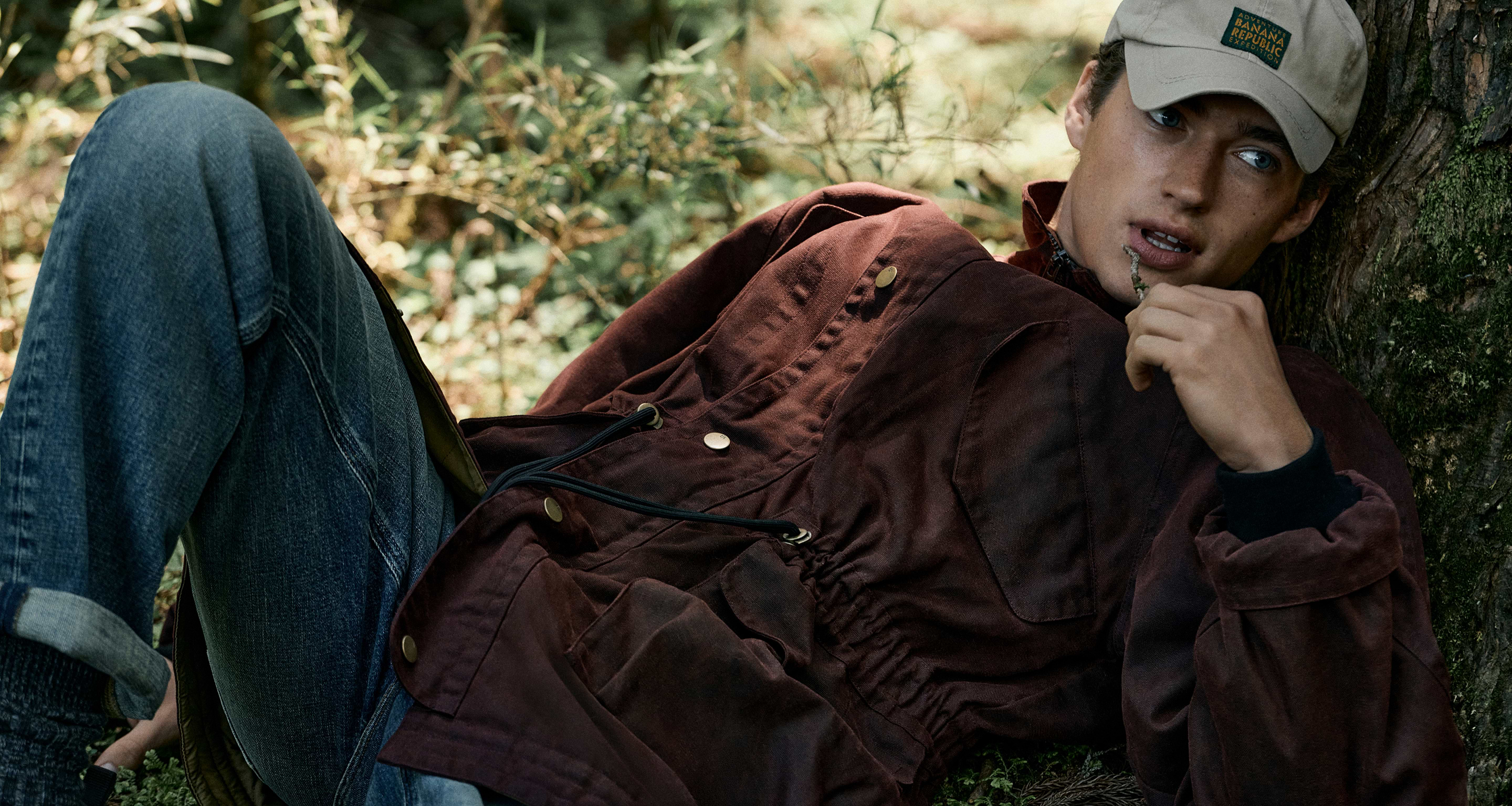 Person in a tan cap, maroon jacket, and jeans reclines against a tree in a forest, holding a twig to their lips.