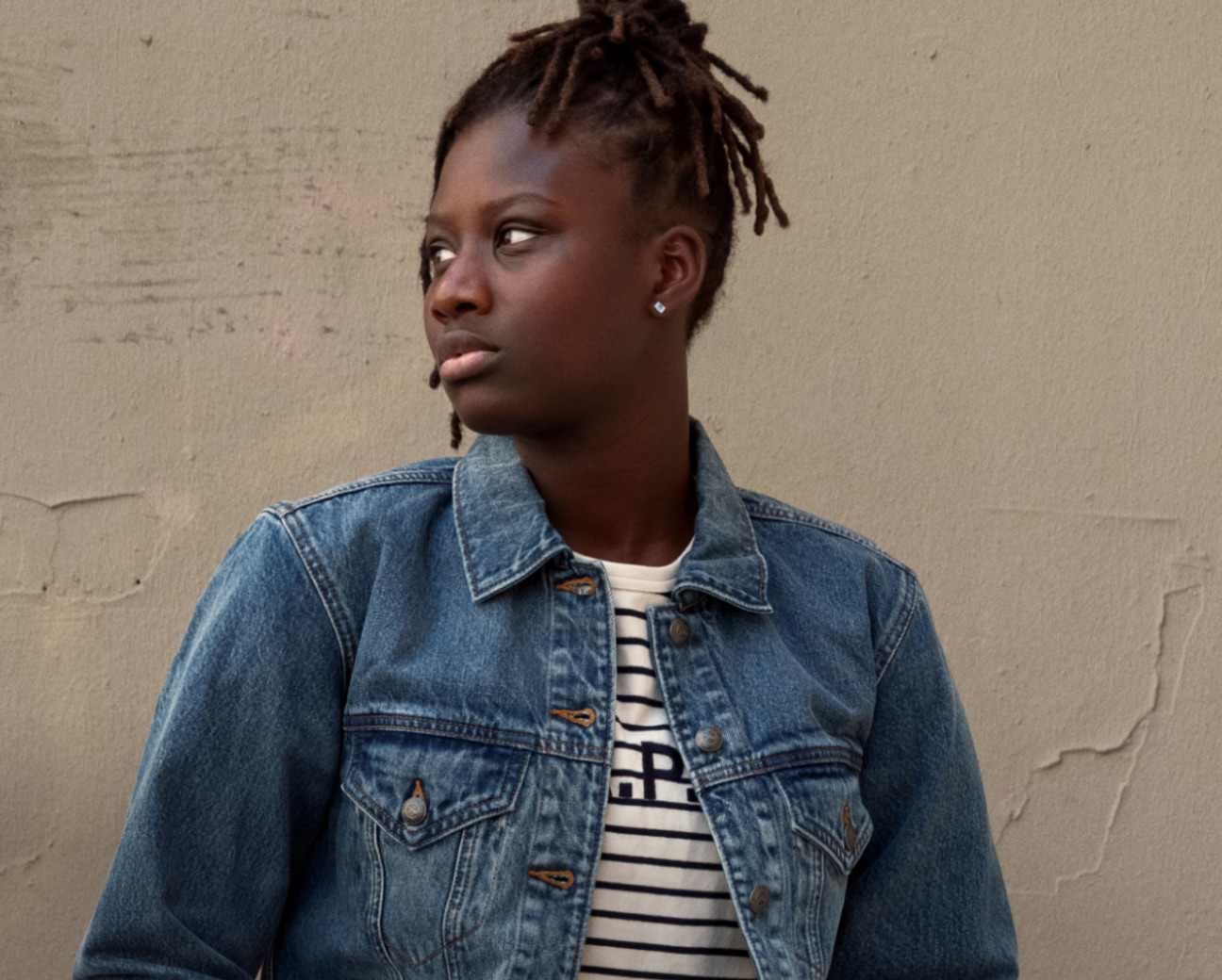 Person with short locs wearing a denim jacket over a striped shirt, looking to the left against a beige wall.