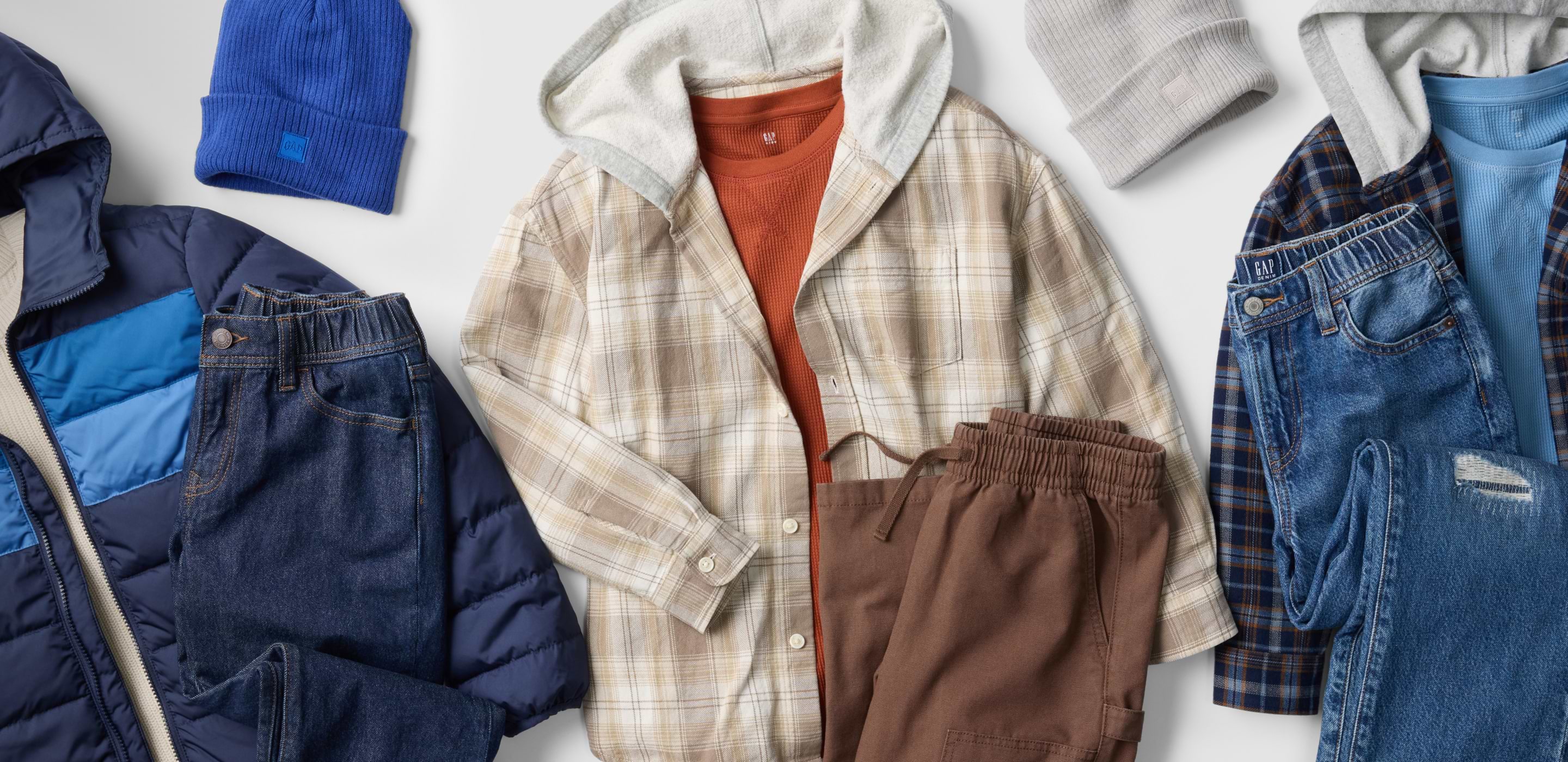 Flat lay of fall clothing: navy puffer jacket, two pairs of jeans, tan and blue plaid hooded shirts over thermal tops, brown joggers, and blue and gray beanies.