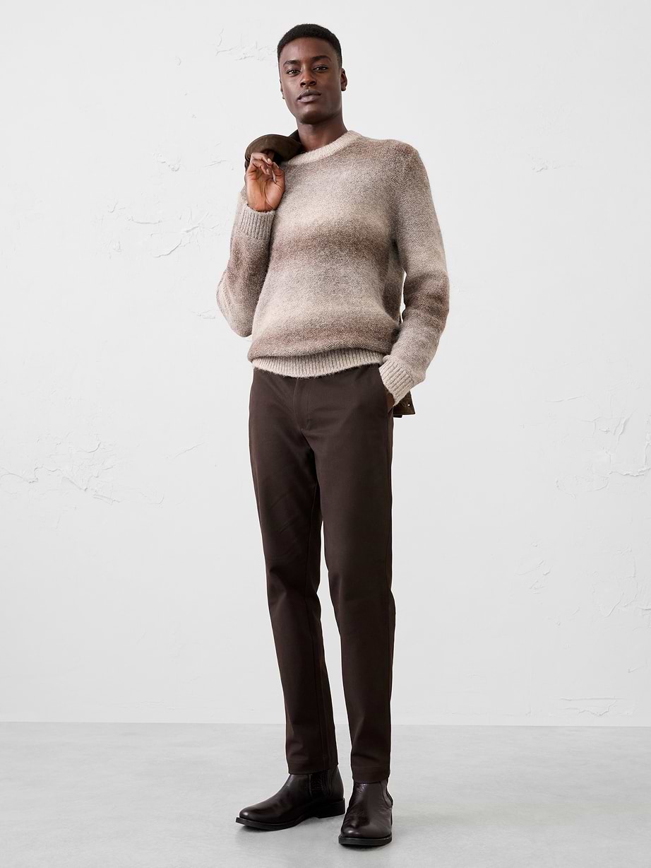 Model wearing a gradient beige-brown sweater, dark brown trousers and boots, holding a coat over one shoulder.