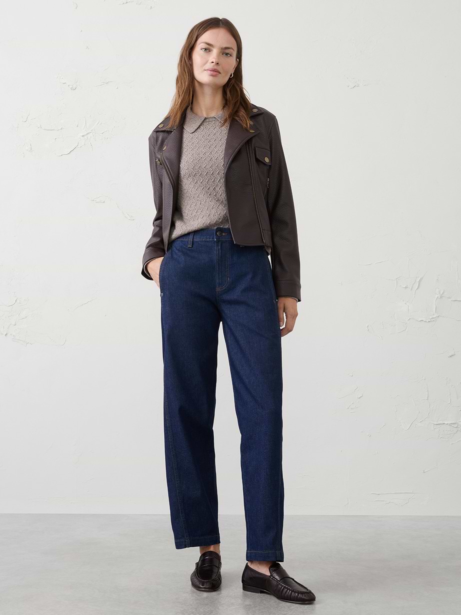 Person standing against a plain light wall wearing a dark leather jacket over a beige knit sweater, dark blue straight‑leg jeans, and black loafers.