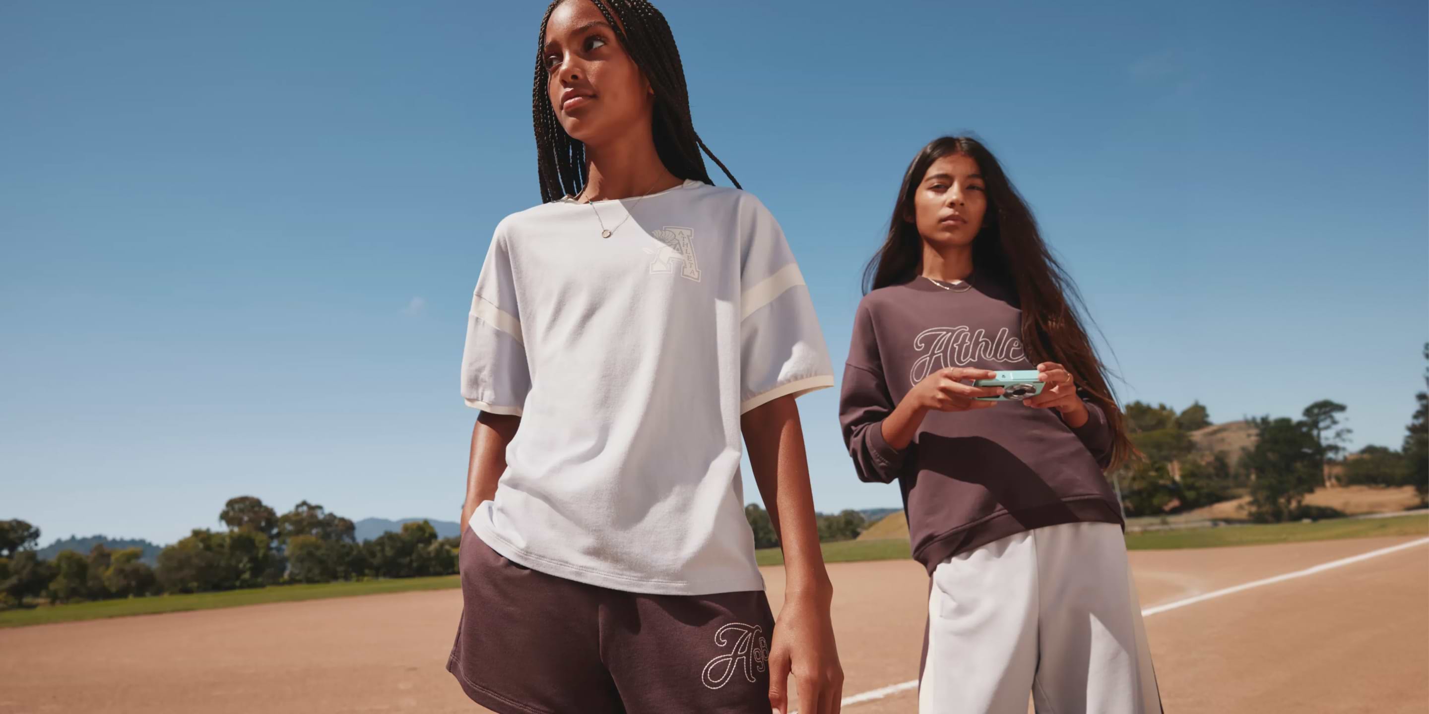 Two girls in sporty outfits stand on a sunny baseball field; one wears a light T‑shirt and shorts, the other a maroon sweatshirt holding a small camera, with blue sky and trees in the background.