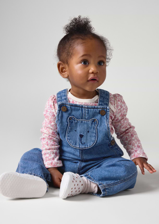 a baby girl sitting on the floor wearing overalls