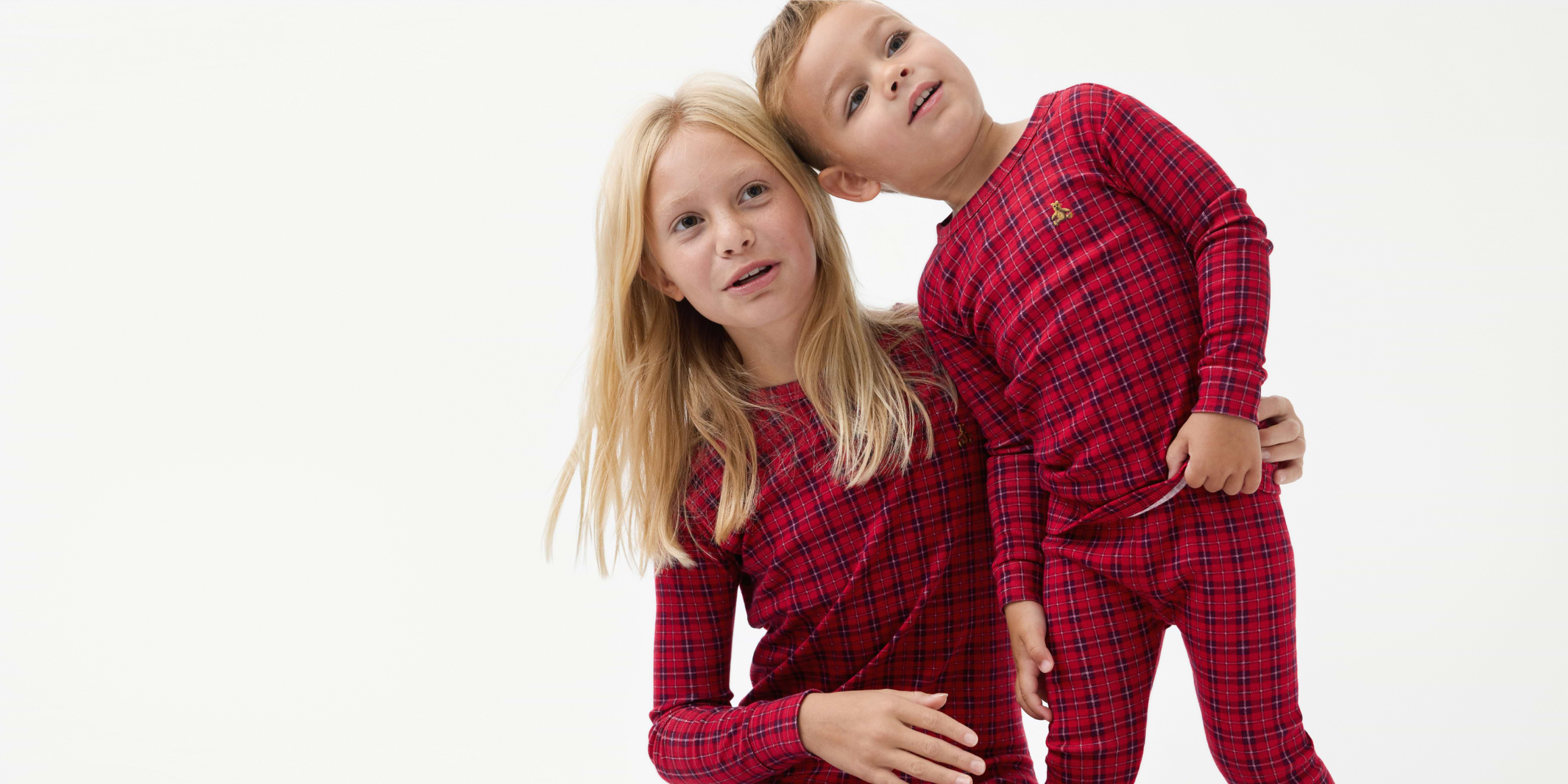 Two children in matching red plaid pajamas lean together and smile against a white background.