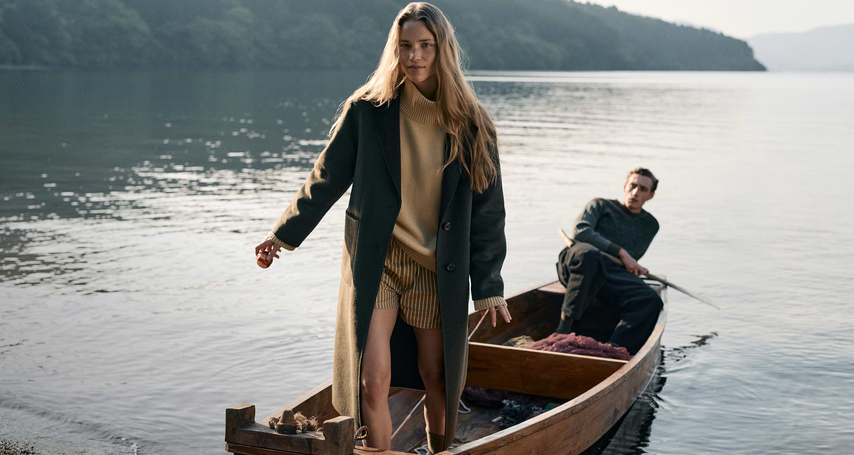 Woman in a long dark coat steps onto a wooden rowboat on a calm lake, with a man rowing behind her.