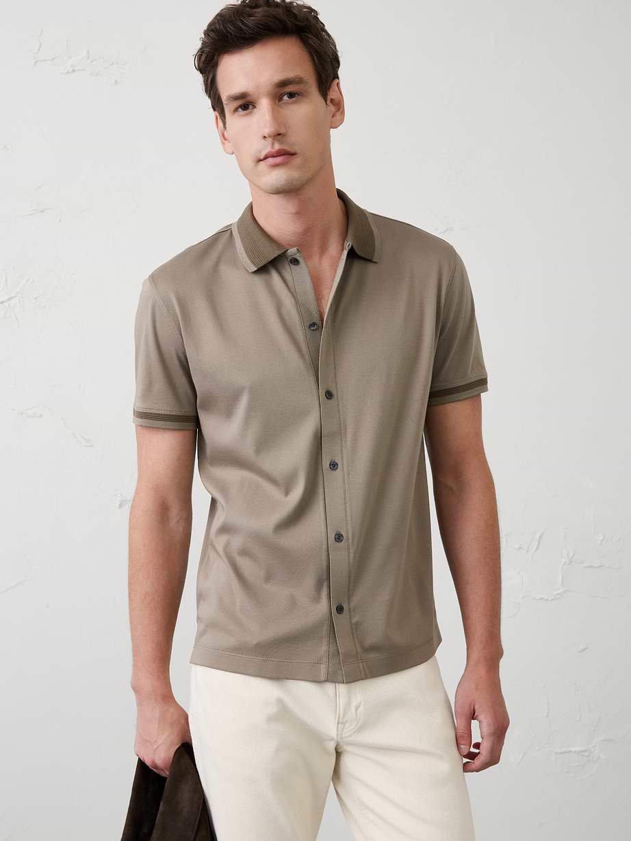 Person wearing a taupe short-sleeve button-front polo and off-white pants, holding a dark jacket against a light textured wall.