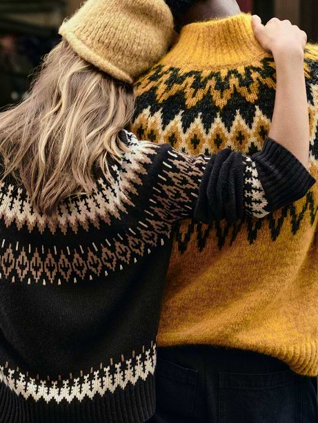 Two people seen from behind in patterned knit sweaters—one black, one mustard—standing close with an arm around the other.
