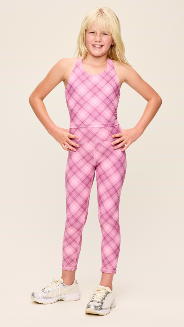 Child in a pink plaid sleeveless jumpsuit and sneakers, smiling with hands on hips in studio.