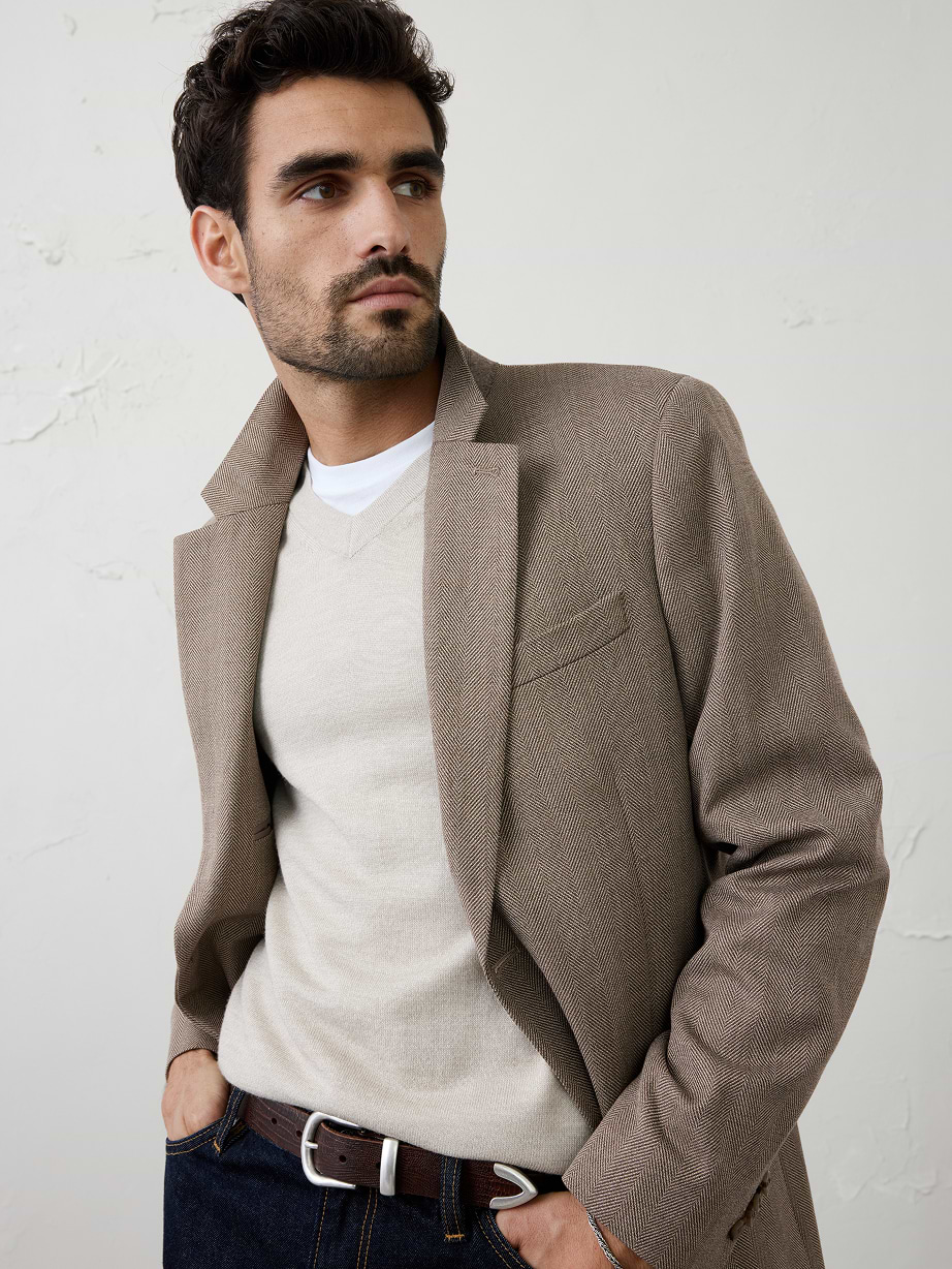 Person in a beige sweater and brown blazer with dark jeans, looking to the side.