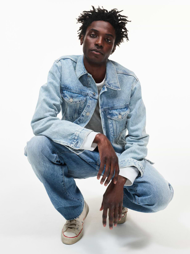 Person crouching in a light-wash denim jacket and jeans with white sneakers against a plain white background.