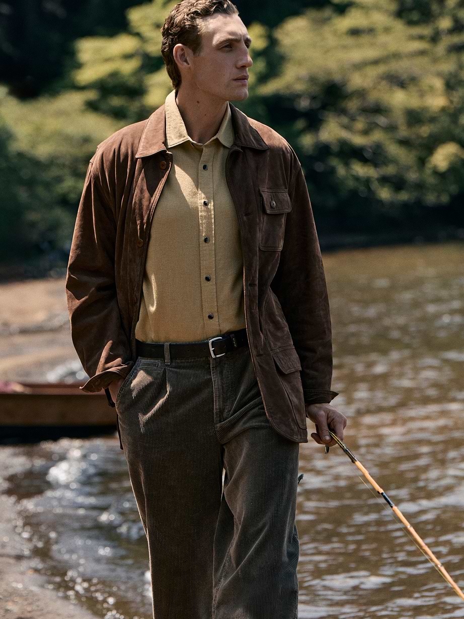 Person in a brown jacket and corduroy pants walking along a riverside, holding a fishing rod, with trees in the background.