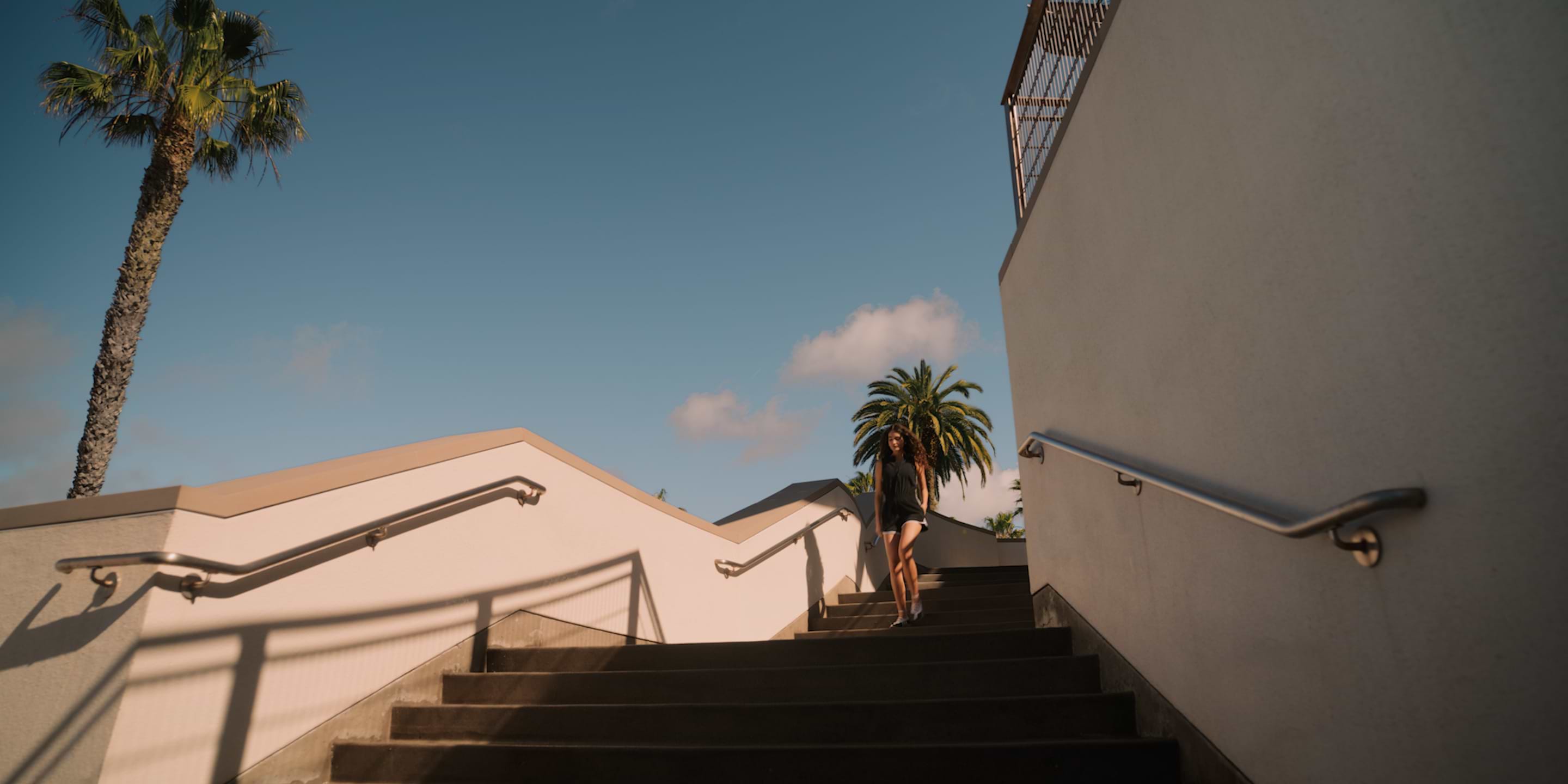 Person walking down sunny outdoor stairs between white walls, with palm trees and blue sky.