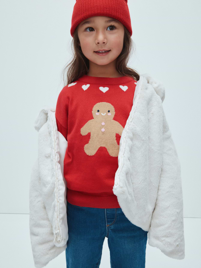 Child in a red beanie and gingerbread-man sweater, blue jeans, with a white fuzzy coat open.