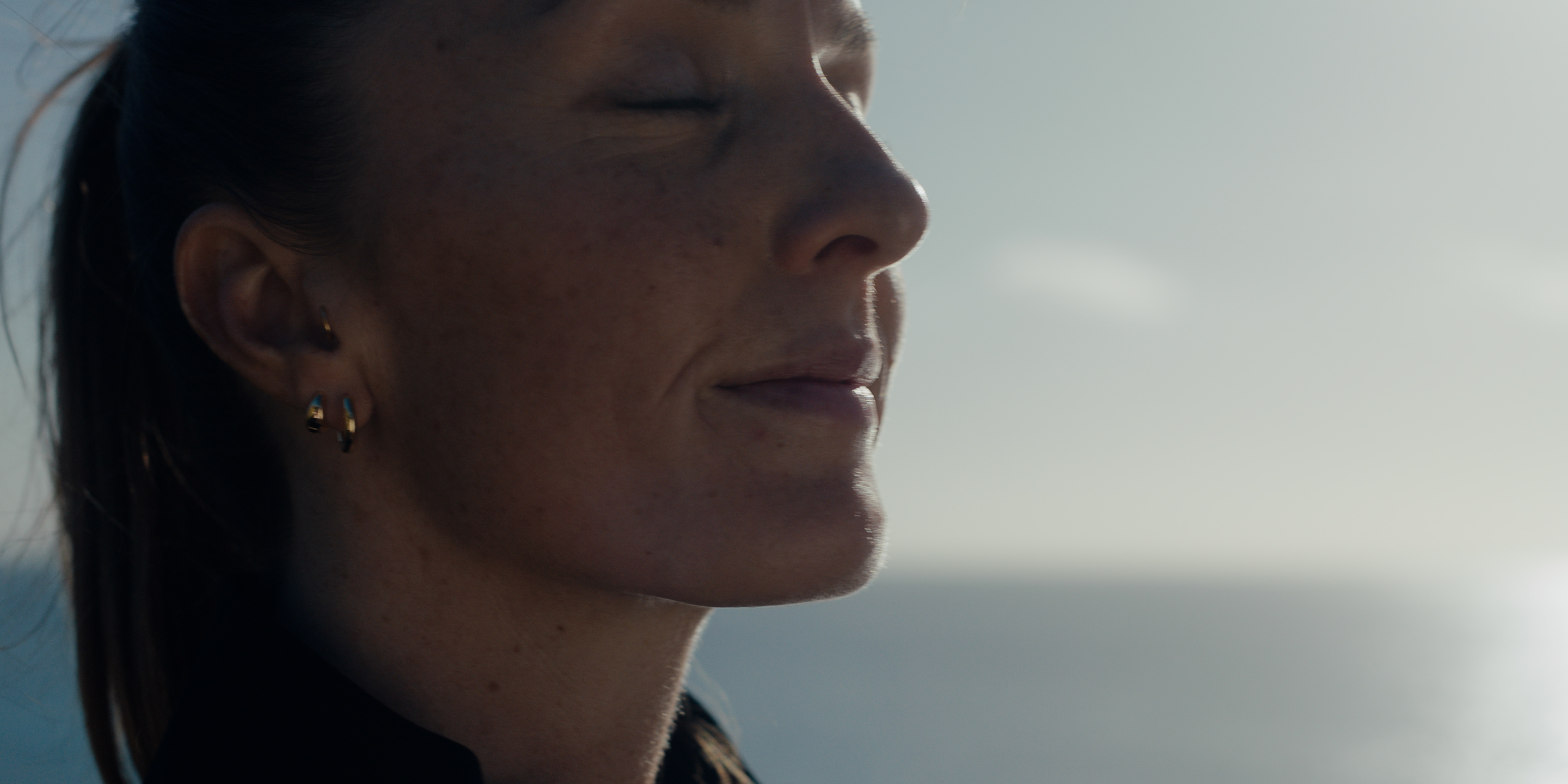 Close-up of an ear with small gold hoop earrings, sunlit ocean behind.