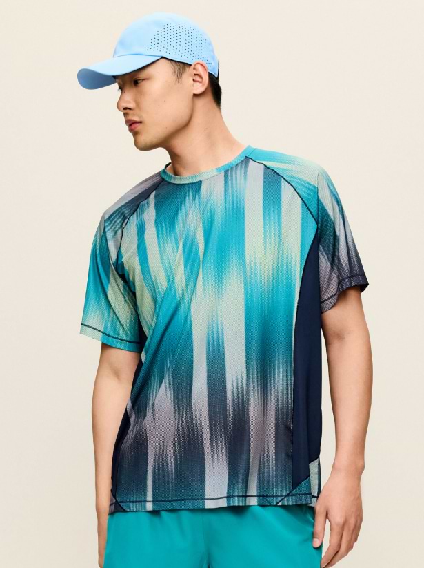 Person in a light blue cap wearing a teal patterned athletic T‑shirt and aqua shorts, looking to the side against a plain background.