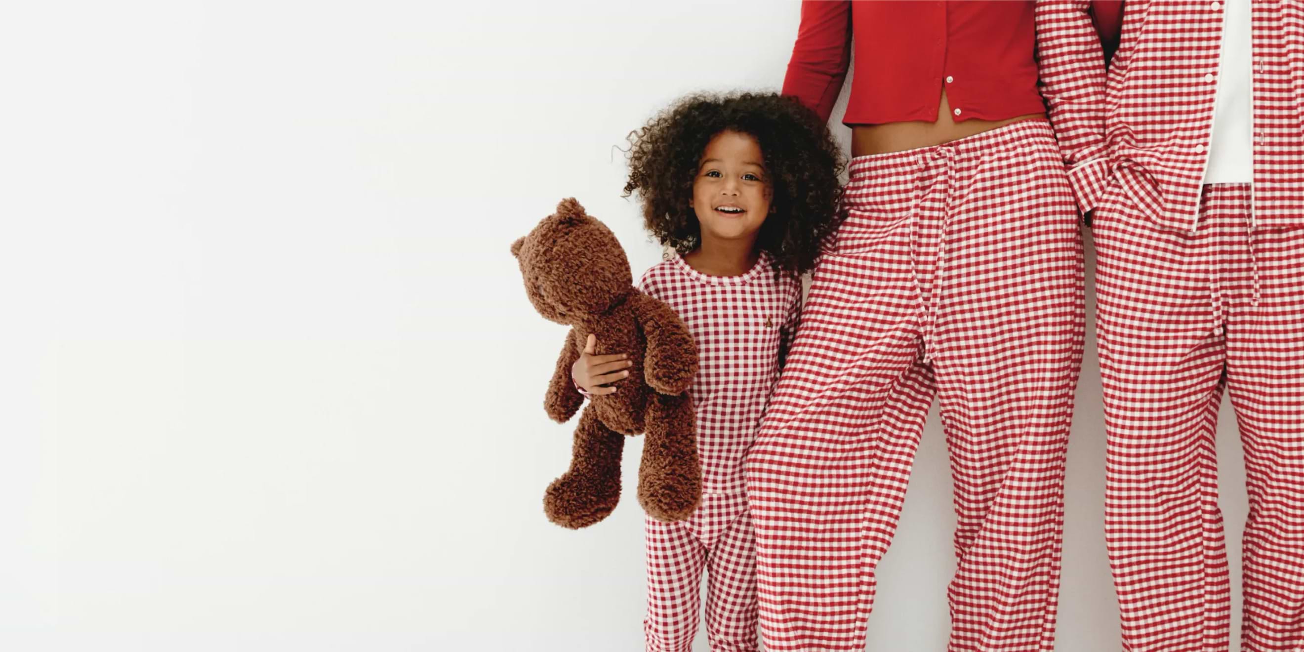 Child holding a teddy bear beside adults in matching red gingham pajamas.