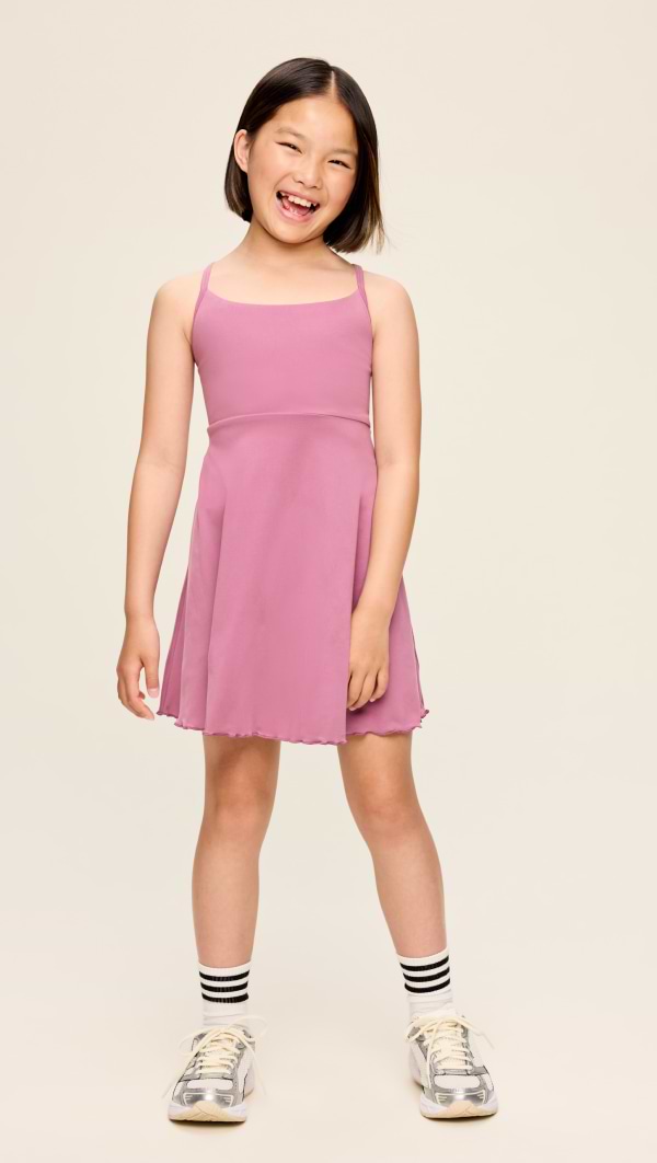 Smiling child in a pink spaghetti-strap dress with striped socks and gray sneakers, standing against a plain beige background.