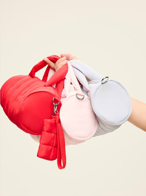 Hand holding red, pink, and light gray puffy mini duffel bags with a red keychain pouch against a plain background.