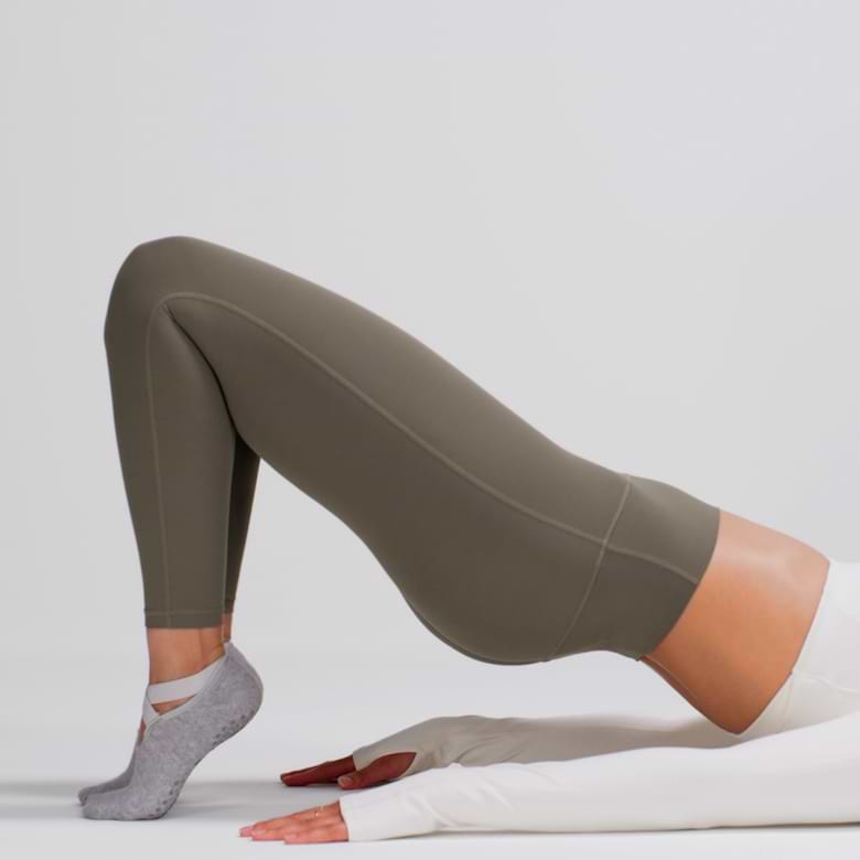 Person in olive leggings and grey grip socks performing a glute bridge, hips lifted with hands on the floor.