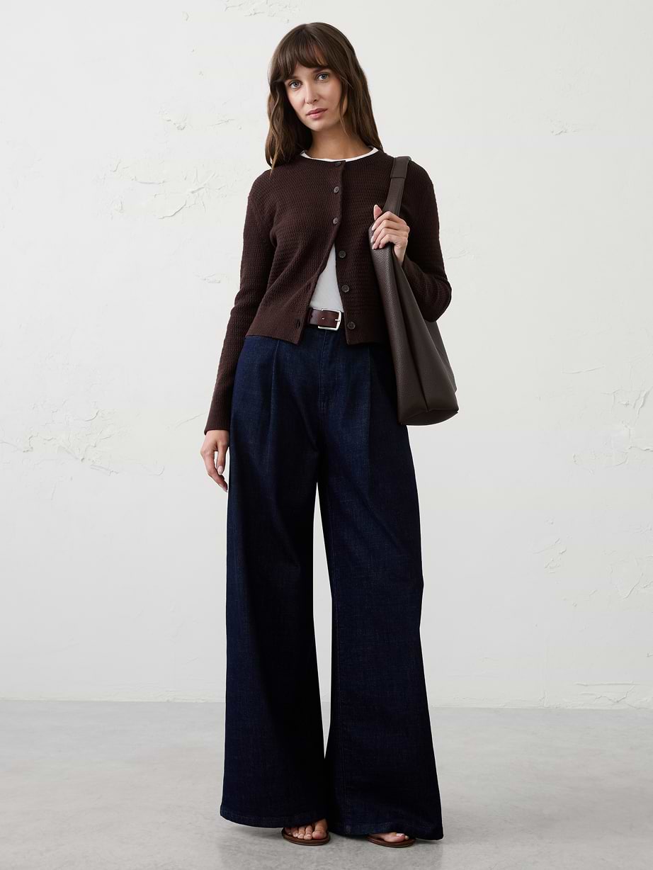 Person in a brown buttoned cardigan over a white tee, wide-leg dark denim trousers, carrying a brown shoulder bag.