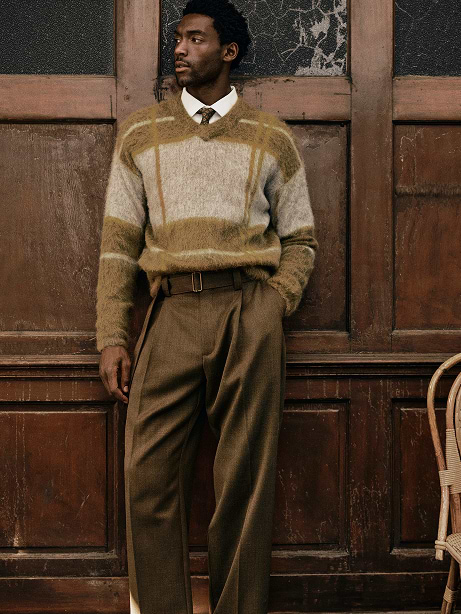 Person in a fuzzy plaid sweater, tie, and pleated brown trousers stands against a vintage wooden paneled wall.