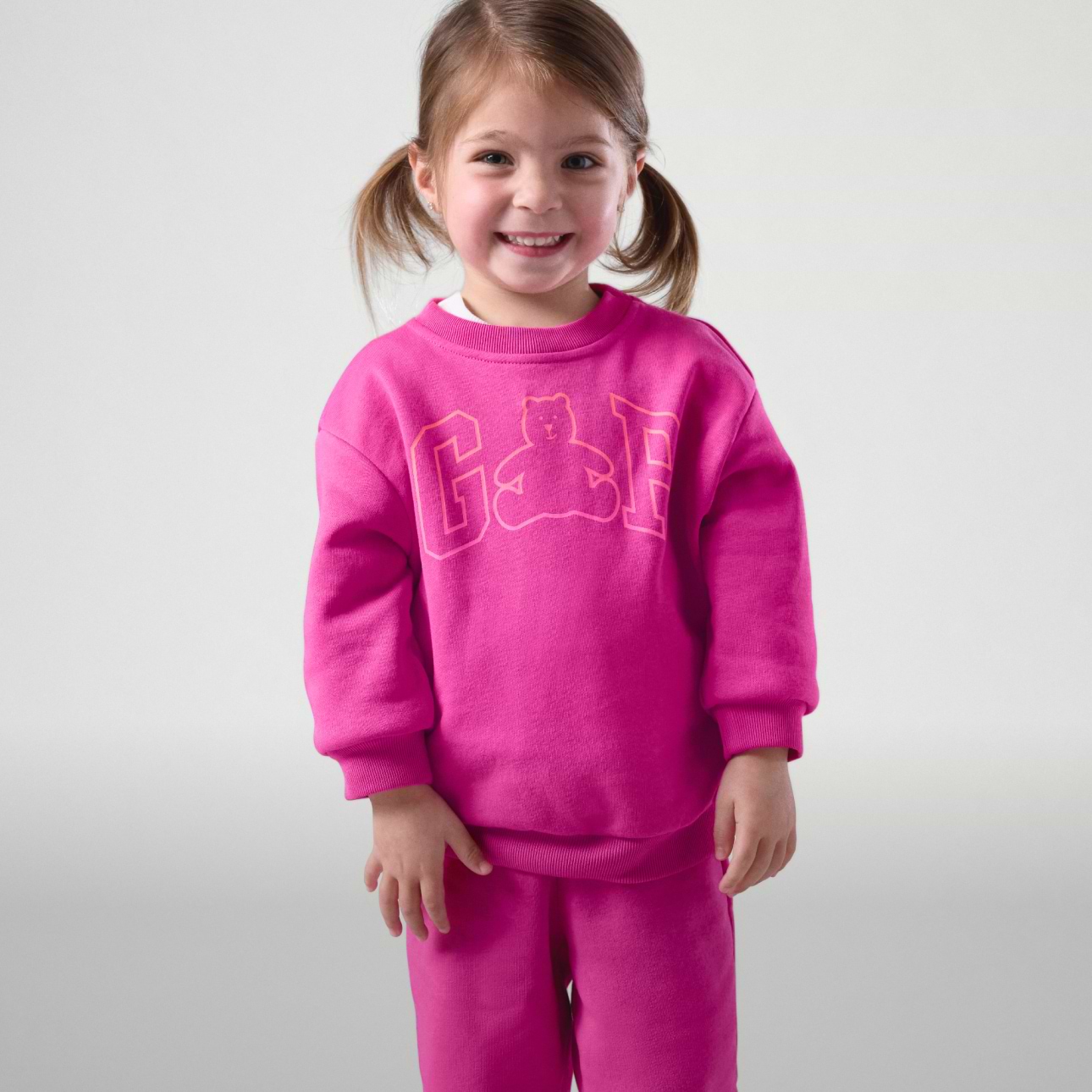 Smiling young child in a bright pink matching sweatsuit with “GAP” and a bear logo, standing against a plain gray background.