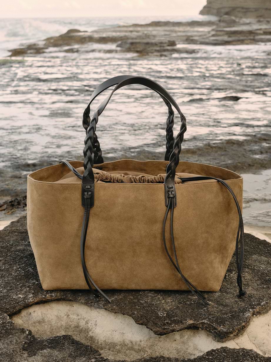 Tan suede tote bag with braided black leather handles and a drawstring top, set on a rocky shoreline by the sea.
