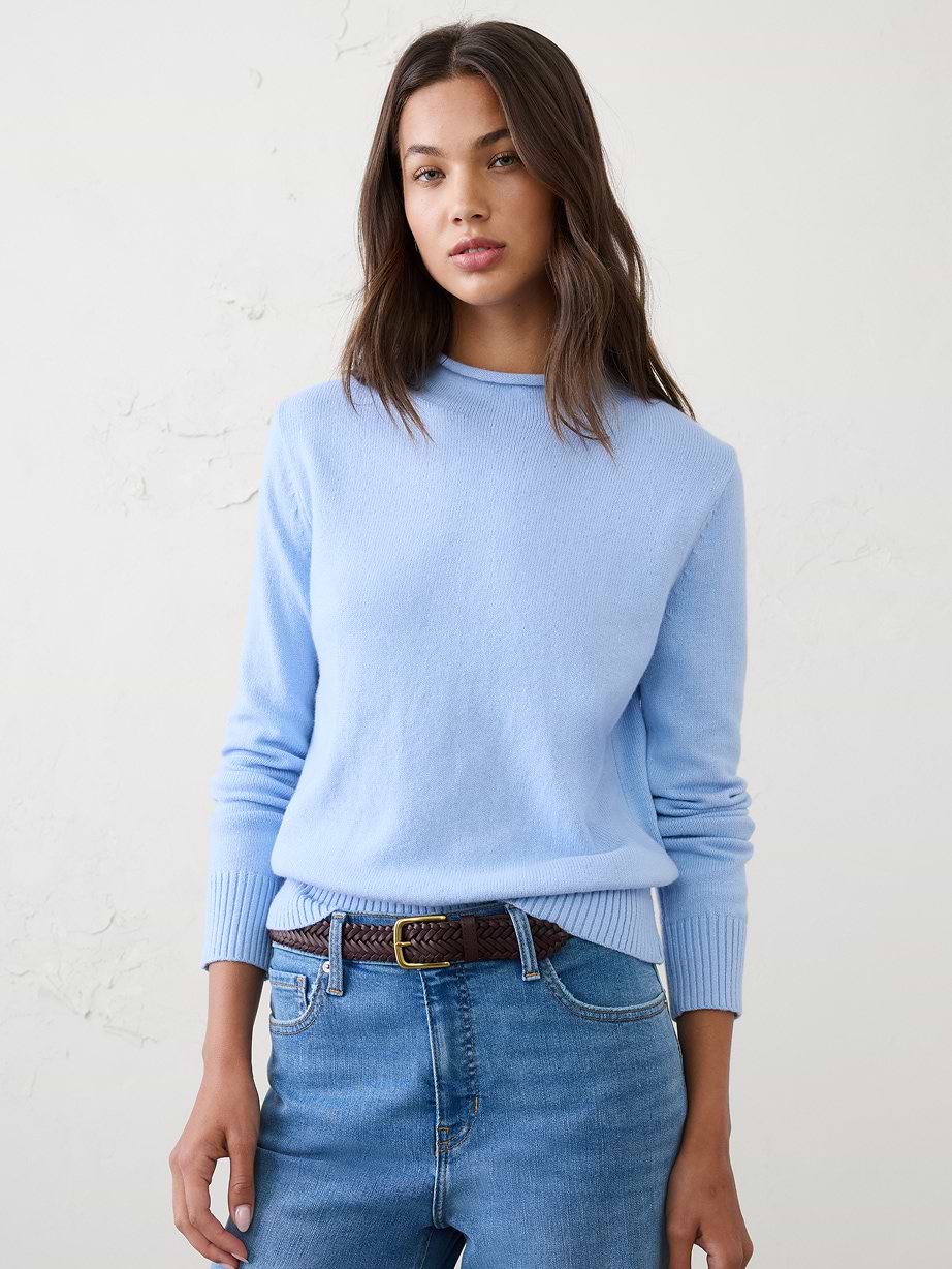 Person wearing a light blue sweater and blue jeans with a brown braided belt, standing against a plain wall.