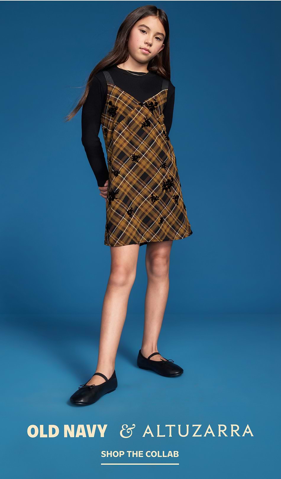 Young model in a brown plaid slip dress over a black long-sleeve and black flats on a blue backdrop; text reads “Old Navy & Altuzarra—Shop the Collab.”