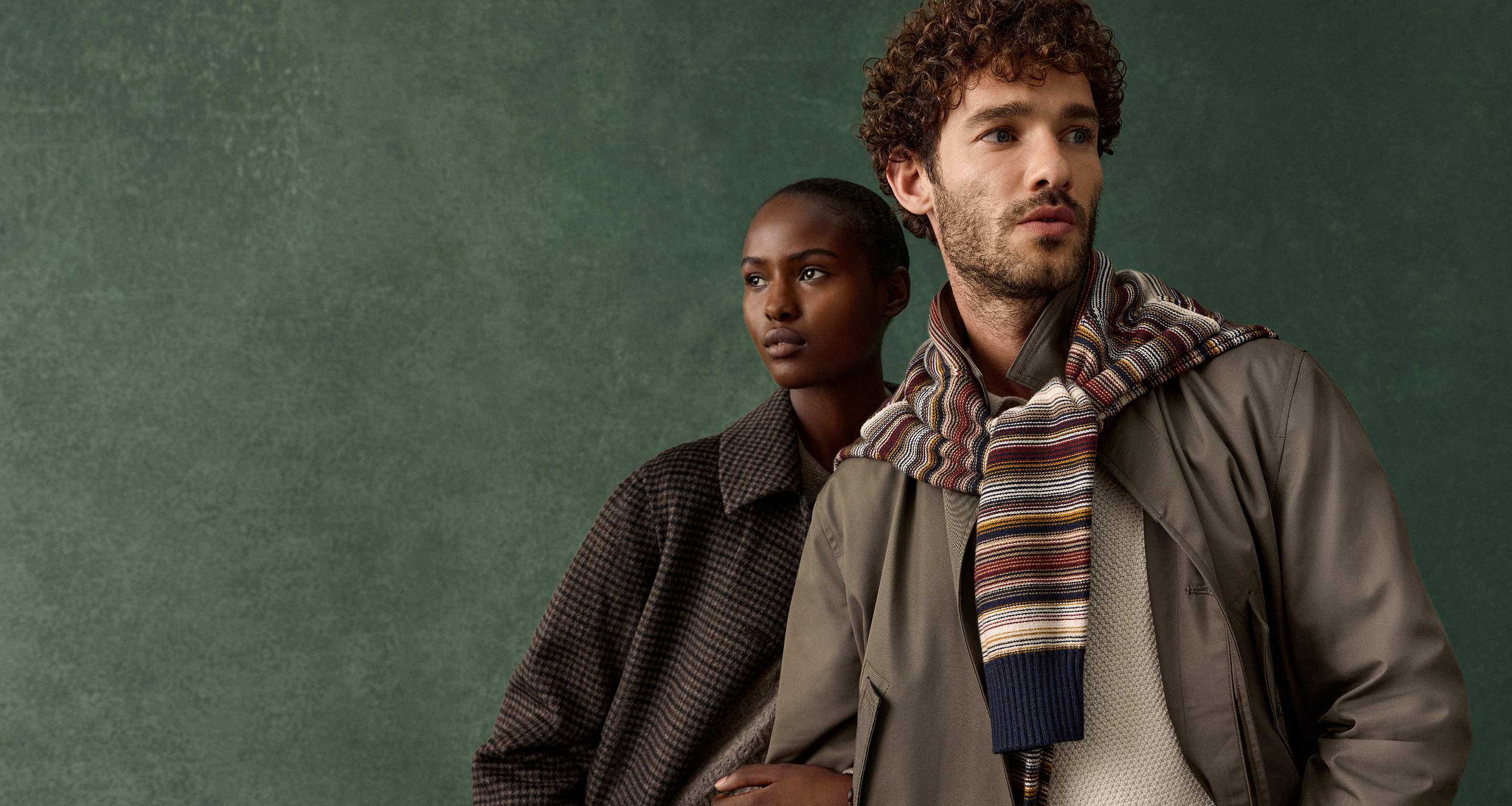 Two models in layered fall outfits—one in a checked coat, the other in a jacket with a striped scarf—against a green backdrop.