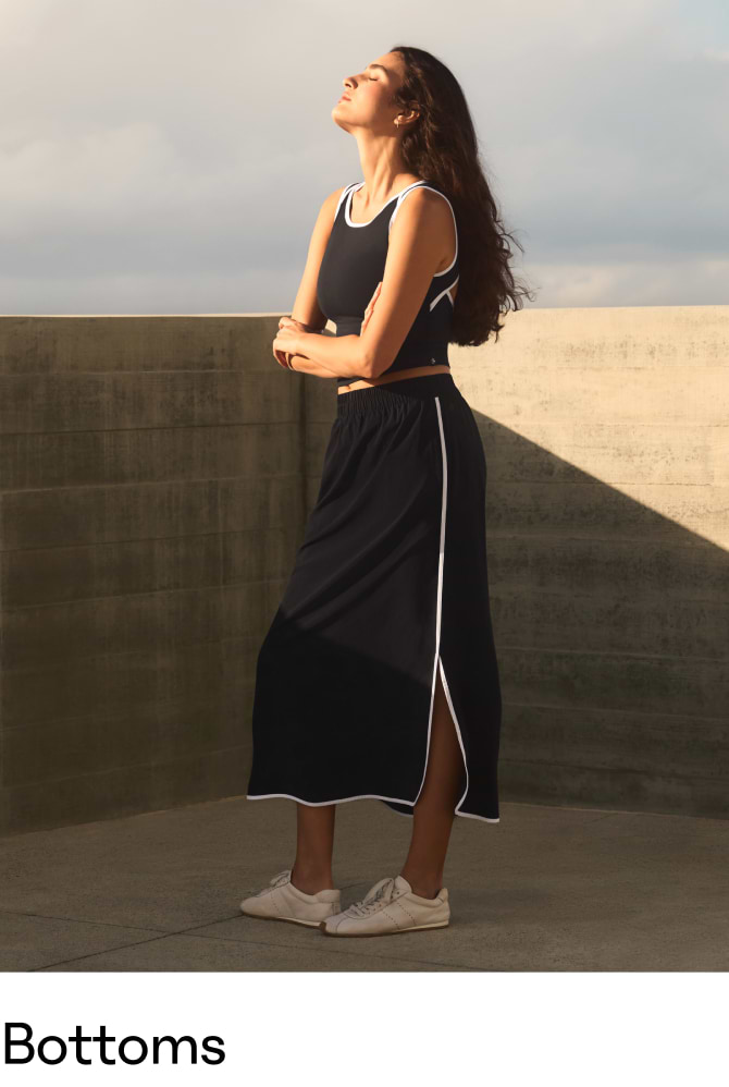 Person standing outdoors in profile, wearing a black midi skirt with white piping and side slit, a matching sleeveless top, and white sneakers.